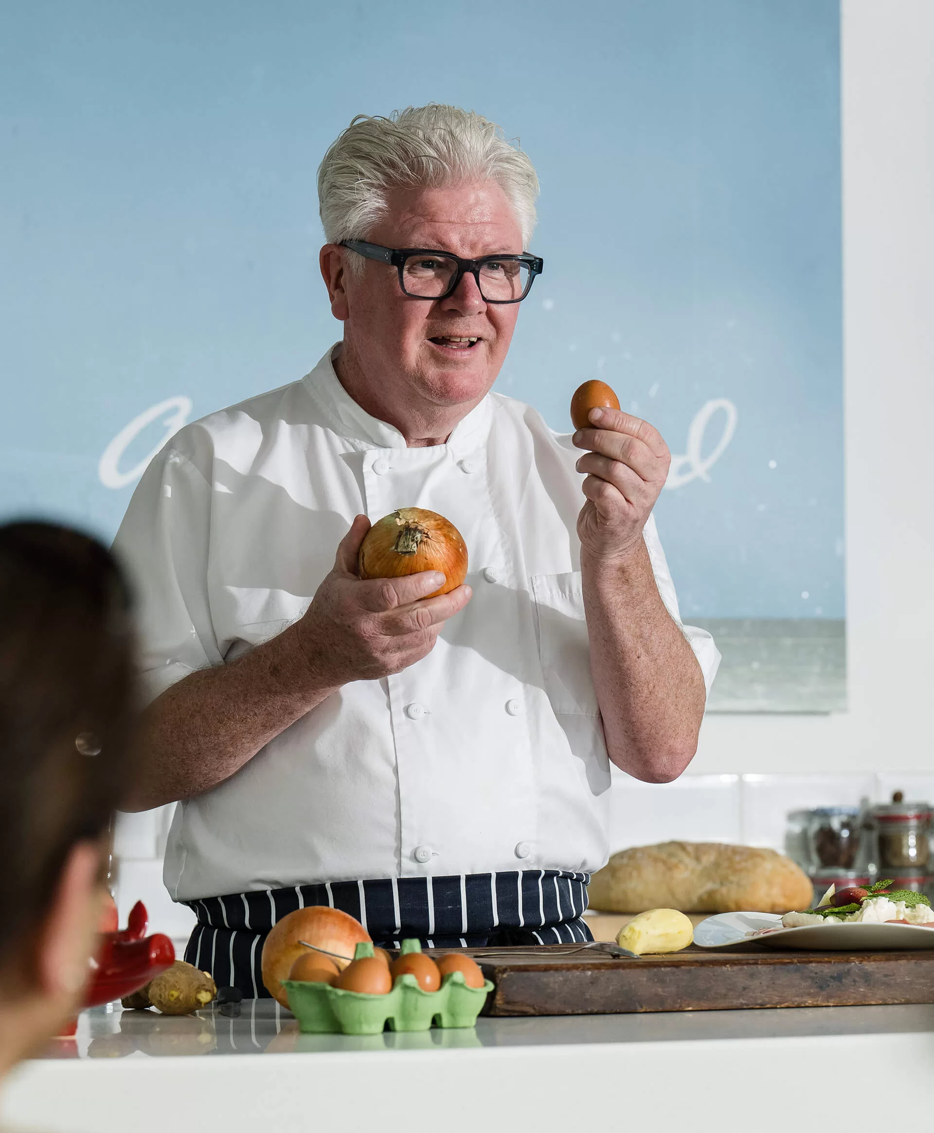 Psul Flynn holding an egg in one hand and an onion in another hand while wearing an apron in front of people