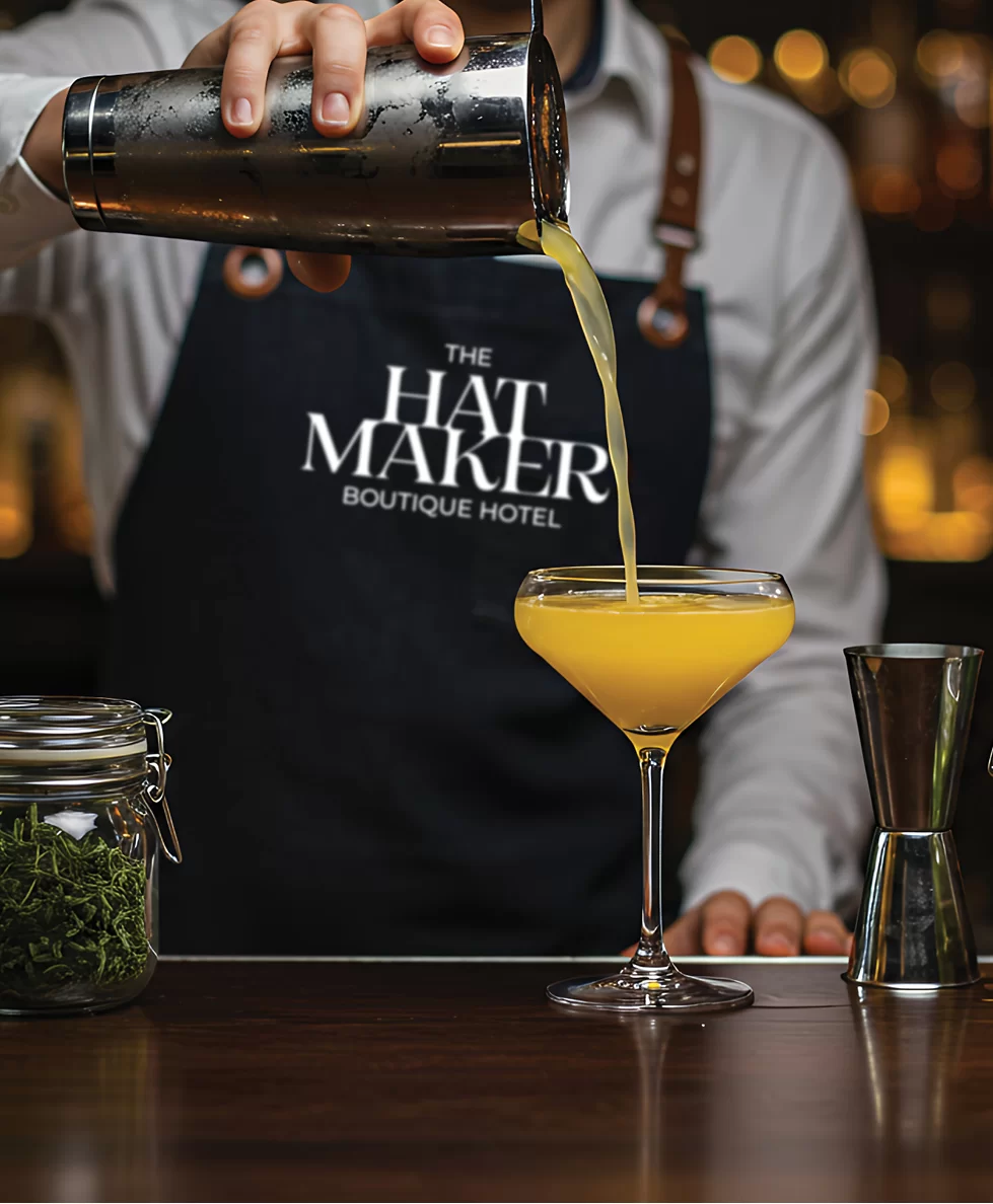 The Hatmaker logo on a barman apron pouring a yellow cocktail from a shaker into a cocktail glass