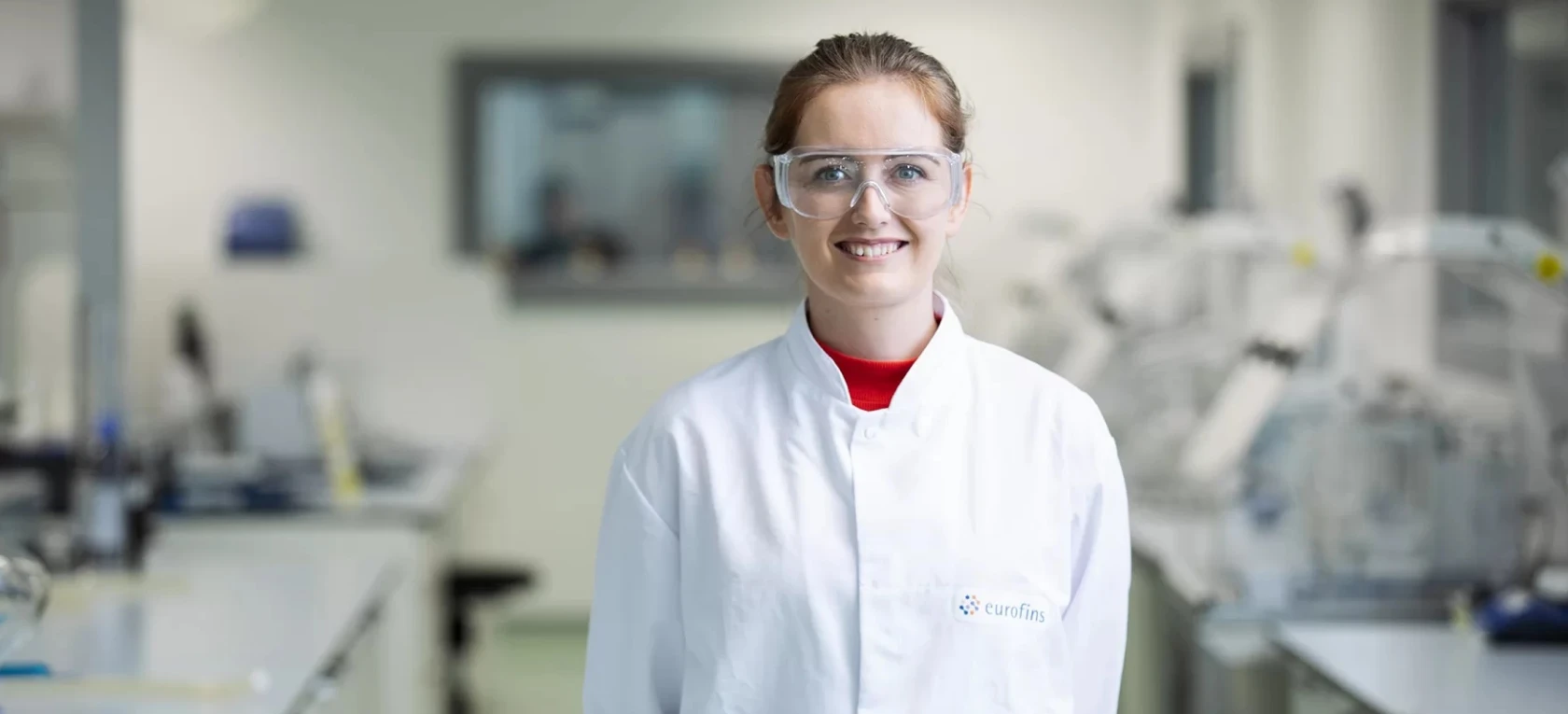 white female Lab worker smiling at camera while wearing protective glasswear and a white coat